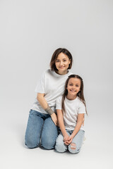 Wall Mural - happy mother with tattoo on hand and short hair hugging preteen brunette daughter while sitting together in white t-shirts and blue denim jeans on grey background, child protection day