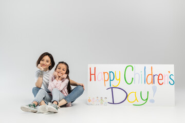 Wall Mural - Full length of smiling tattooed mother looking at camera while sitting near daughter and placard with happy children's day lettering during celebration on grey background