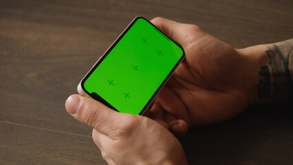 Focus on Chroma Display and hands. 4k footage. Caucasian man using smartphone in horizontal mode with a green mock-up screen is lying on the wooden floor inside and watching videos or movies.