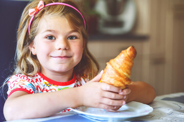 Wall Mural - Smiling child at breakfast. Food and happy kids. The girl is eating a croissant. Cute preschool girl having healthy meal.