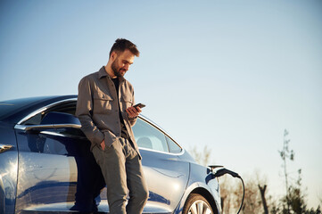 Wall Mural - Holding smartphone in hand. Man is standing near his electric car outdoors