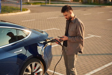 Wall Mural - Bored, waiting for automobile to charge. With smartphone. Man is standing near his electric car outdoors