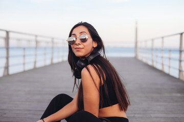 Young woman in sunglasses and black headphones enjoying music, sitting by the river side, on a bridge sunset light 