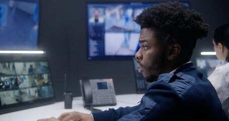 African American officer controls CCTV cameras with AI face scanning in police monitoring room. Female operator works at background. Big screens on the wall showing security cameras footage. Close up.