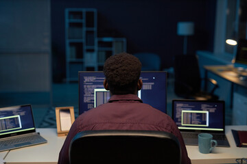 rear view of programmer sitting at his workplace in front of computers and working till late night