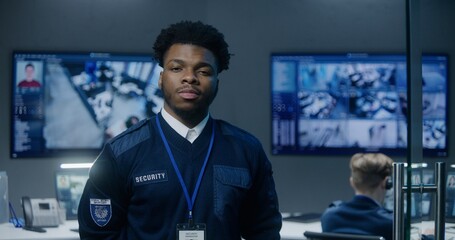 Portrait of African American security officer in police monitoring center looking at camera. Employees work on computers on background. Big digital screen with surveillance CCTV cameras video footage.