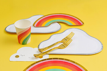 Wall Mural - Cute paper party plates in the shape of rainbow and clouds with cups for themed kids party. Birthday party decorations on yellow background. Set of holiday disposable tableware for party or picnic.