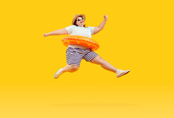 Full body photo of a happy funny fat plus size overweight woman in sunglasses with rubber ring jumping and having fun on studio yellow background. Summer holiday trip and vacation concept.
