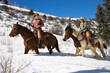 Man and young woman riding horses in deep snow across a country landscape. Horizontal shot.