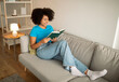 © Prostock-studio - Happy pretty millennial curly black woman reading book, enjoy free time and rest at weekend on sofa