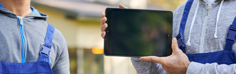 Cropped shot of young male builder showing screen of digital tablet pc with coworker holding blueprint in the background, working at cottage construction site