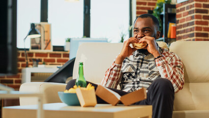 African american man taking bite of hamburger with fries, eating takeaway meal from delivery at home. Happy person enjoying fast food and drinking bottle of beer, feeling relaxed. Handheld shot.