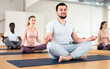 © JackF - Young smiling man sitting in lotus pose on mat in fitness room, doing yoga meditation with group of adult people..