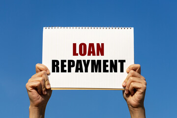 Loan repayment text on notebook paper held by 2 hands with isolated blue sky background. This message can be used as business concept about loan repayment.