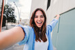 © Jose Calsina - Happy young caucasian student lady looking at camera and taking a selfie portrait having fun, standing outside. Front view of laughing woman shooting a photo for social media at the university campus