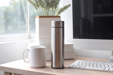 Blank mockup white coffee mug and steel thermo bottle for water on office table near computer and window