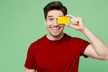 Poster - Young smiling happy man he wear red t-shirt casual clothes hold in hand cover eye with mock up of credit bank card isolated on plain pastel light green background studio portrait. Lifestyle concept