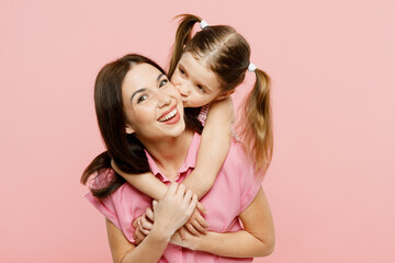 Wall Mural - Happy woman wear casual clothes with child kid girl 6-7 years old. Daughter stand behind mother hug and cuddle, looking camera, kiss isolated on plain pastel pink background Family parent day concept
