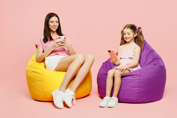 Wall Mural - Full body woman wear casual clothes with child kid girl 6-7 years old. Mother daughter sit in bag chair hold in hand use mobile cell phone isolated on plain pink background. Family parent day concept.