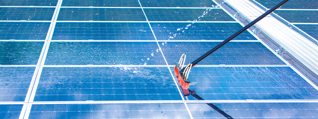 Professional worker cleaning solar panels with brush and washing with water on roof structure of building factory. Technician using mop to clean the dirty and dust, green electricity energy technology