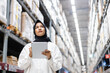 © eakgrungenerd - Asian muslim female headscarf hijab holding digital tablet checklist product shelves warehouse. woman worker pointing finger inspecting goods industry. Logistic concept.
