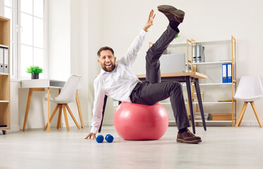 Funny, joyful businessman having sports workout in office. Portrait of happy, smiling, energetic man sitting on red fit ball and raising one leg up. Fitness, exercise, healthy lifestyle concept