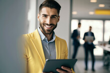 Generative AI Image Of Smiling Young Male In Yellow Blazer Looking At Camera While Standing With Tablet In Hand In Office Against Blurred Window With Colleagues