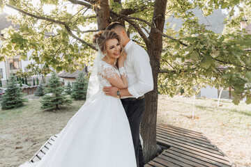 Wall Mural - Wedding portrait. A groom in a black suit and a blonde bride are standing, hugging and kissing under a tree. Photo session in nature. A beautiful ray of the sun in the photo. Beautiful hair and makeup