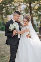 Wall Mural - Wedding portrait. The groom in a black suit and the blonde bride are standing, hugging, posing holding a bouquet under a tree. Photo session in nature. Beautiful hair and makeup