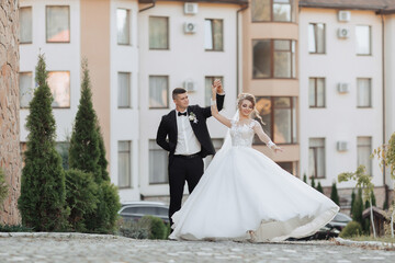 Wall Mural - Wedding portrait. A groom in a black suit and a blonde bride dance a waltz against the background of buildings. Long dress in the air. Photo session in nature. Beautiful hair and makeup