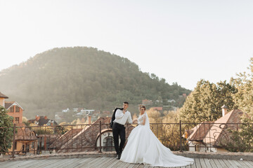 Wall Mural - Wedding portrait. The groom in a black suit and the blonde bride are standing, hugging, posing holding a bouquet under a tree. Photo session in nature. Beautiful hair and makeup