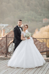 Wall Mural - Wedding portrait. The groom in a black suit and the blonde bride are standing, hugging, posing holding a bouquet under a tree. Photo session in nature. Beautiful hair and makeup