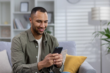 A young African American man is sitting on the couch at home and using the phone. Writes a message to a girl, chats with friends, reads news, social networks. Close-up photo