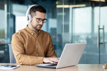 A young man sits at a workplace in the office, works, studies on a laptop with headphones. Listens to the webinar, music, course