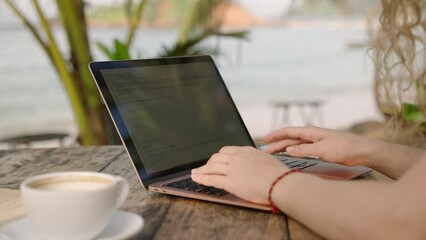 Young female developer working on laptop by the ocean. Woman freelancer coding at outdoor tropical cafe. Caucasian girl working remotely typing on computer at exotic location. Close-up sideview shot