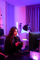 Asian pretty gamer girl use joystick when playing video game on computer screen while sitting ergonomic chair in entertainment pink neon room