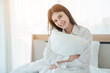 © kelvn - Photo of young happy woman in pajama stretching her arms and smiling while sitting on bed after sleep