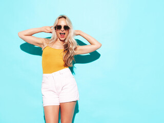 Wall Mural - Young beautiful smiling blond female in trendy summer shorts clothes. Sexy carefree woman posing near blue wall in studio. Positive model having fun. Cheerful and happy. Going crazy