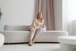 © TrueFrame Collective - Attractive mature Caucasian woman wearing stylish casual outfit sitting relaxed on sofa in spacious living room looking at camera