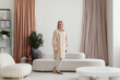 © TrueFrame Collective - Long shot of senior Caucasian woman wearing oversized long shirt standing in modern living room at home looking at camera