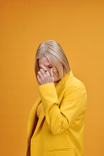 Vertical Medium Studio Portrait Of Mature Caucasian Woman With Blond Hair Wearing Yellow Outfit Posing On Camera Covering Her Face With Hand, Bright Yellow Background