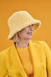 © Clique Images - Vertical medium close-up portrait of modern mature Caucasian woman wearing stylish yellow outfit with hat looking away, bright yellow background