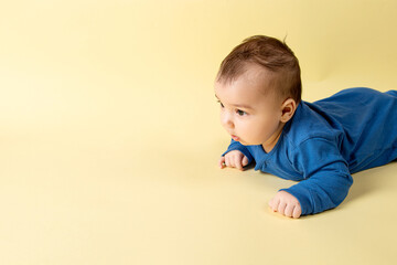 Wall Mural - A beautiful little baby in a blue bodysuit, lies on the stomach on a yellow background. Concept, mockup, blank, copy space.