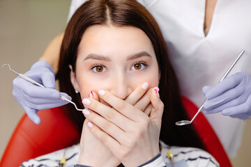 Closeup shot of a terrified female covering her mouth with both hands at the dentist appointment. Her nightmare