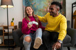 © Studio Marmellata - Cheerful multiracial boyfriend and girlfriend in colorful casual clothes sitting on sofa with gamepads and playing video game together in modern living room