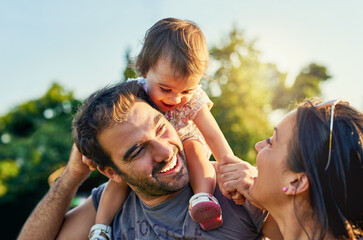 Family, father and daughter on shoulders in park with happy mom, love and summer sunshine. Young couple, baby girl or laugh together for freedom, bond and helping hand for care, backyard or garden