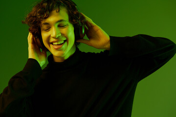 Wall Mural - Man teenager wearing headphones listening to music, dancing and singing with his eyes closed, DJ party happiness and smile, hipster lifestyle, portrait green background mixed neon light, copy space