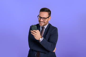 Wall Mural - Smiling handsome business manager text messaging over smart phone while standing against blue background. Portrait of cheerful male professional checking social media applications over cellphone