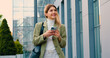 © serg - Cheerful young caucasian woman walking in the street to the office with cellphone in hand. Smiling lady holding cellphone in hands outside