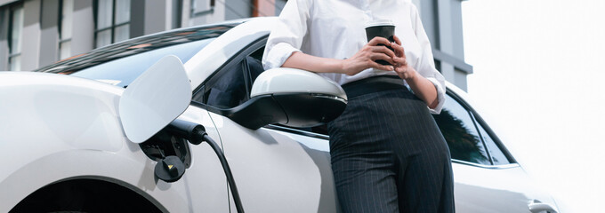 Closeup progressive suit-clad businesswoman with her electric vehicle recharge her car on public charging station in modern city with power cable plug and renewable energy-powered electric vehicle.